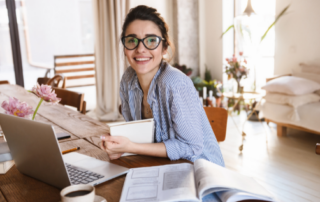 the best 485 Visa Australia lawyer Brisbane - image of lady smiling at the camera sitting at a timber table with laptop, note book and textbook with flowers on the table - call HCM Legal today